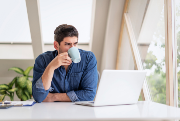 Man in a denim shirt sitting at a desk, drinking from a mug while looking thoughtfully at a laptop screen in a bright, modern room with large windows and indoor plants.